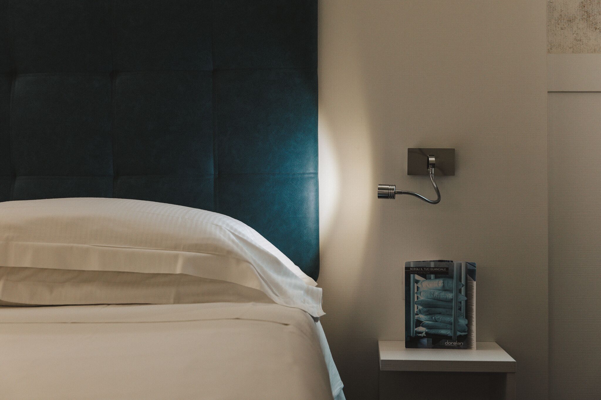 Una cama bien hecha con sábanas blancas, cabecera oscura, mesita de noche con un libro, y una lámpara de pared.