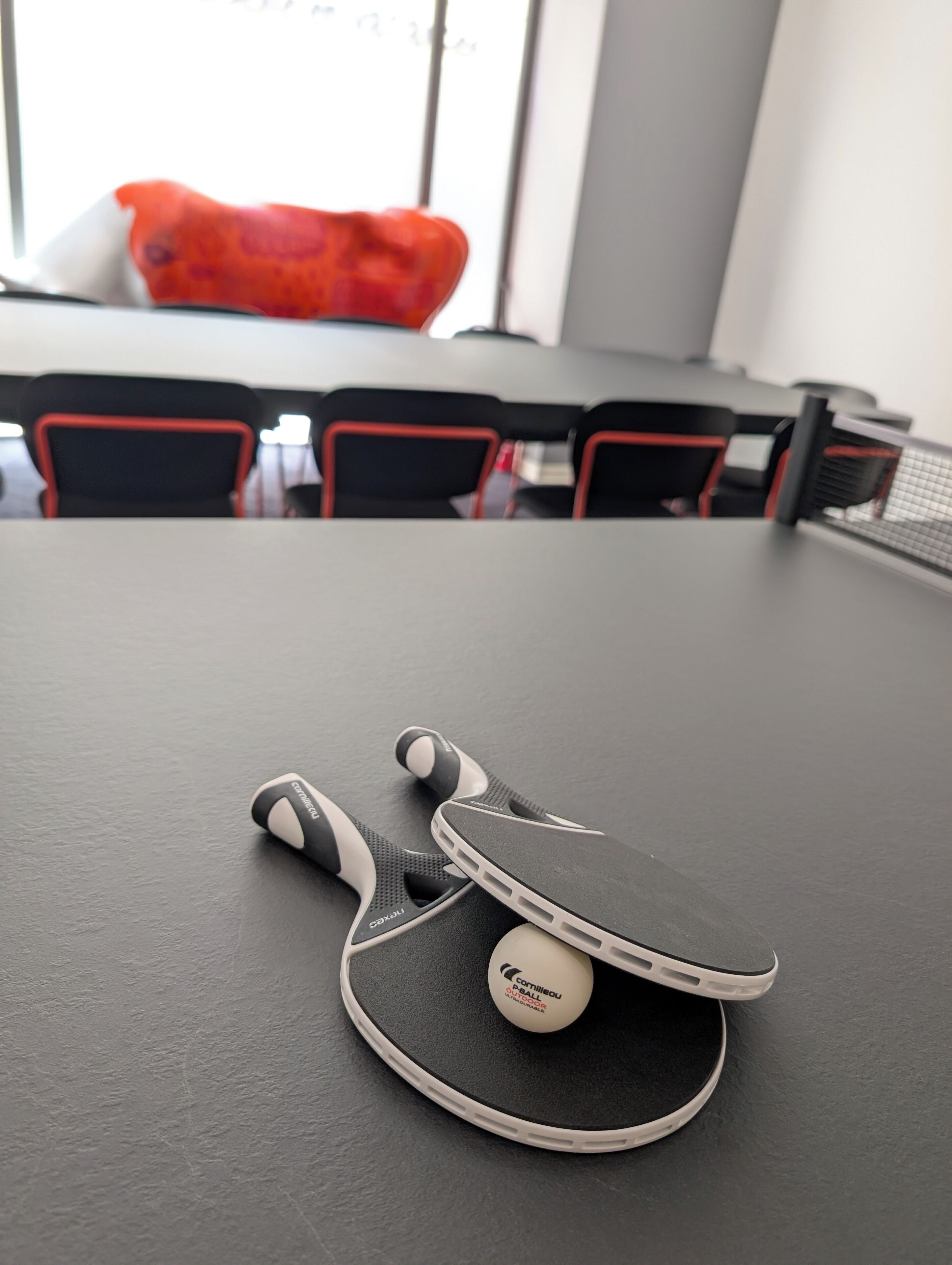 Mesa com uma raquete de tênis de mesa e bola, um travesseiro vermelho e cadeiras pretas com detalhes vermelhos em um quarto com janela.