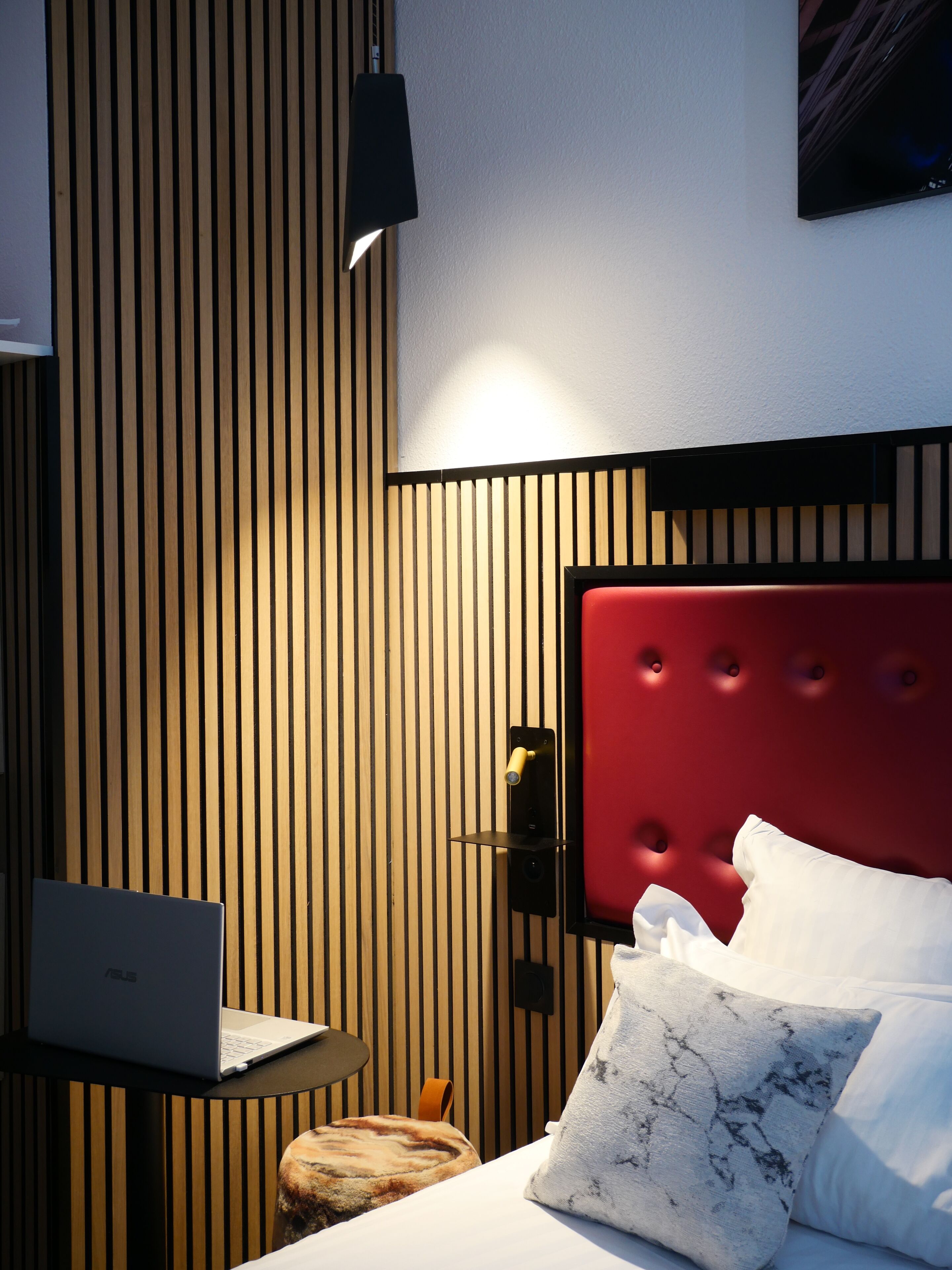 Una habitación de hotel moderna con pared de listones de madera, cabecera tapizada en rojo, un ordenador portátil sobre una mesita pequeña y una cama con ropa blanca y almohada estampada con diseño de mármol.