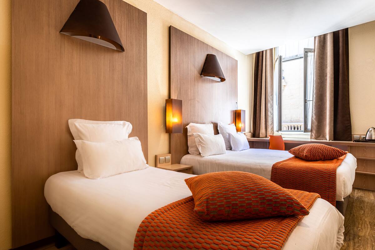 Ein Hotelzimmer mit zwei Betten, jeweils mit weißer Bettwäsche und orangen Zierkissen. An den Wänden sind Lampen angebracht, und ein Fenster mit Vorhängen ist zu sehen.