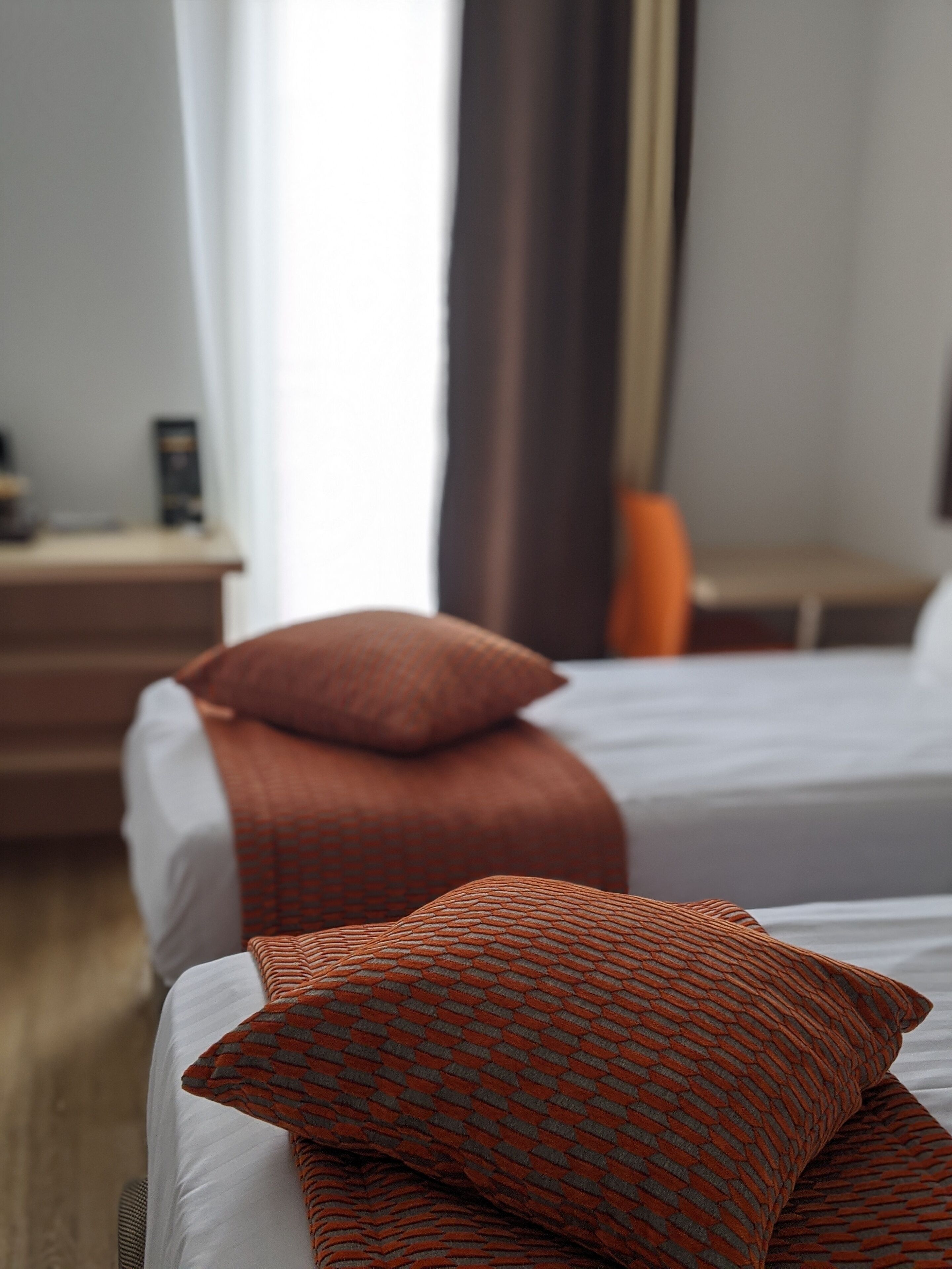 Ein Hotelzimmer mit zwei Betten, jedes mit einem orangefarbenen, gemusterten Kopfkissen. An der Seite steht ein Nachttisch mit einer Lampe und ein kleiner Tisch mit einem Buch.