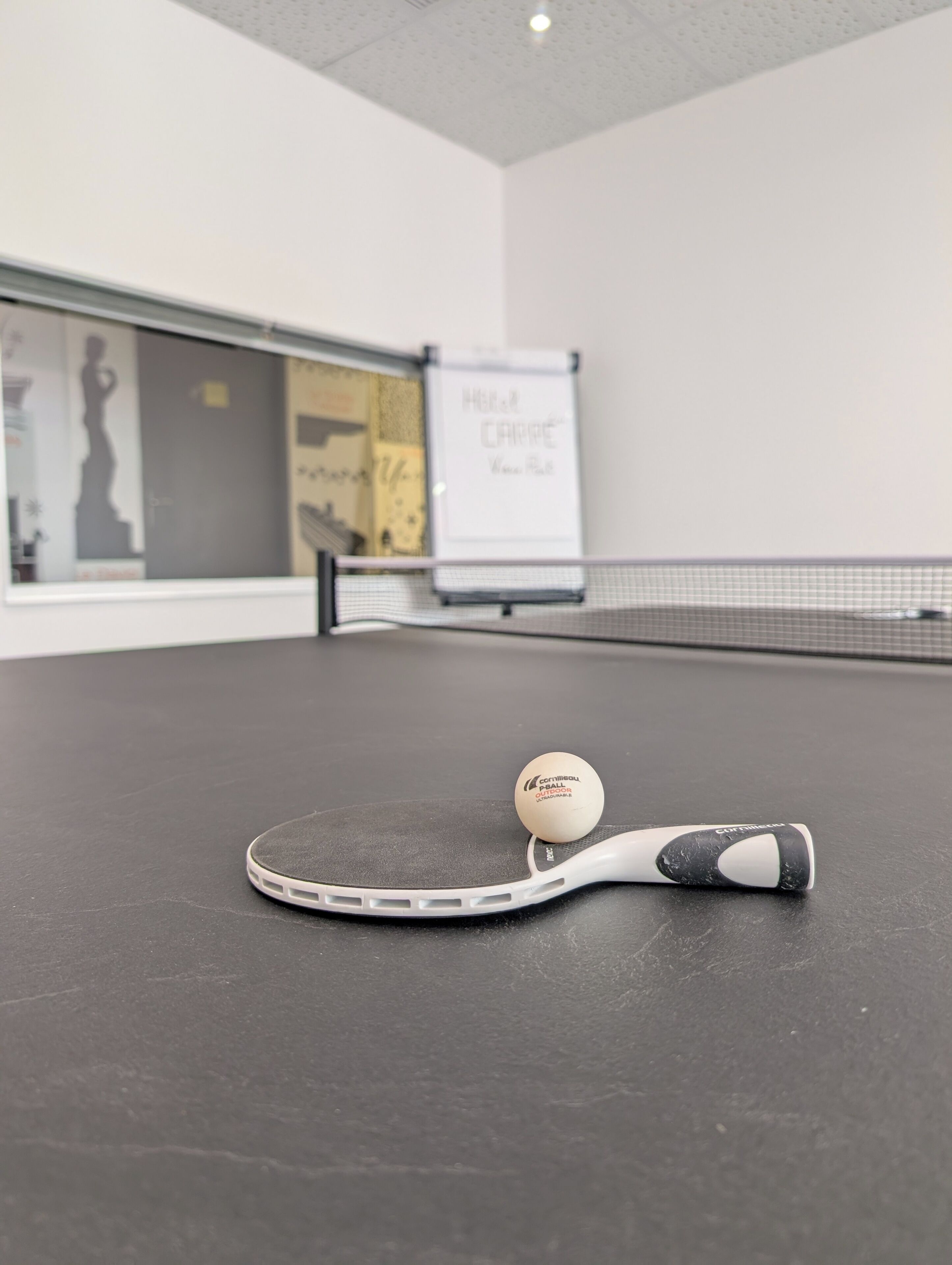 Uma mesa de pingue-pongue com uma bola e uma raquete em um espaço com um quadro branco e cartazes na parede.