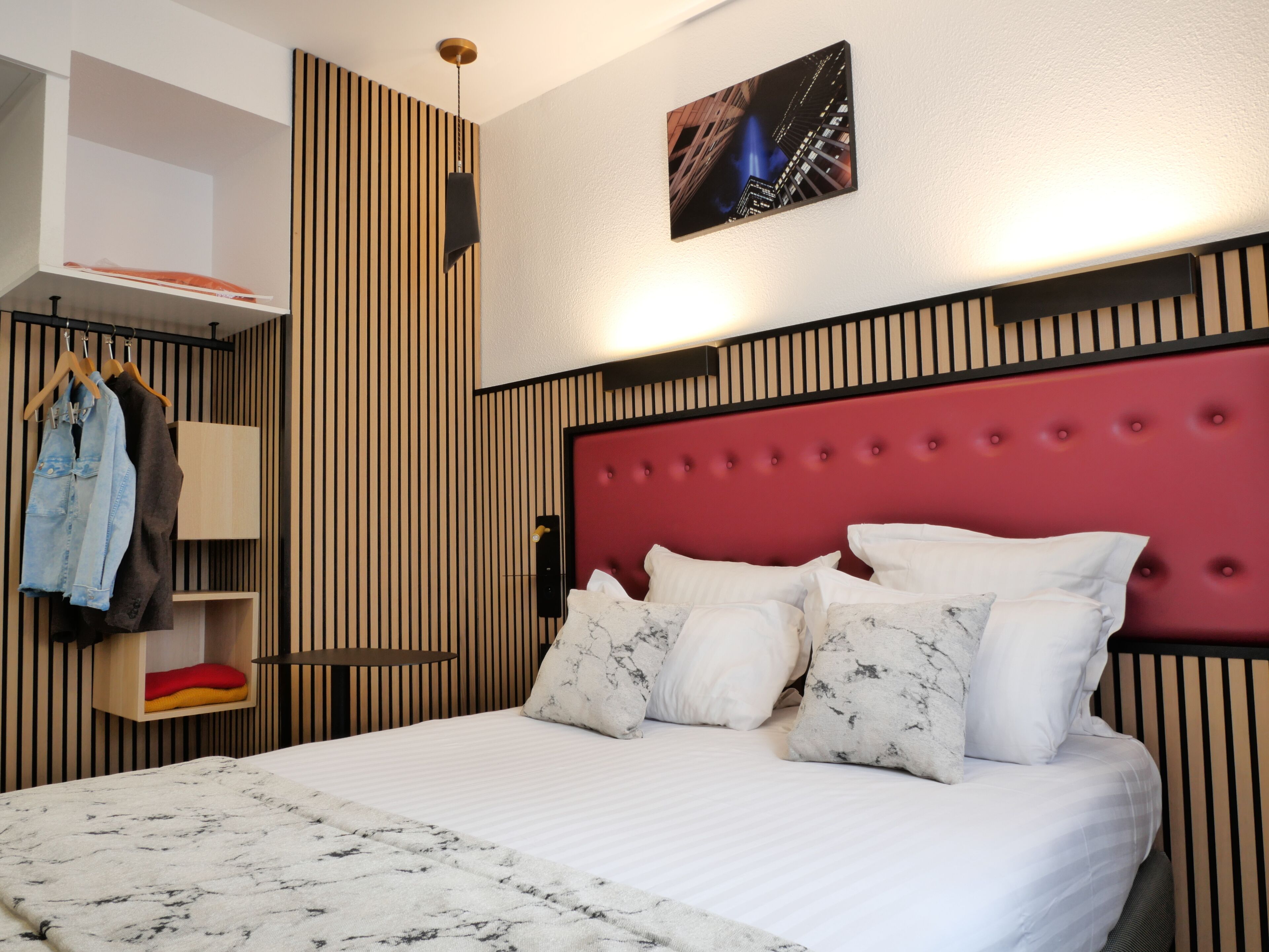 A hotel room with a bed, a red headboard, a wall-mounted shelf with clothes, and a painting above the bed.