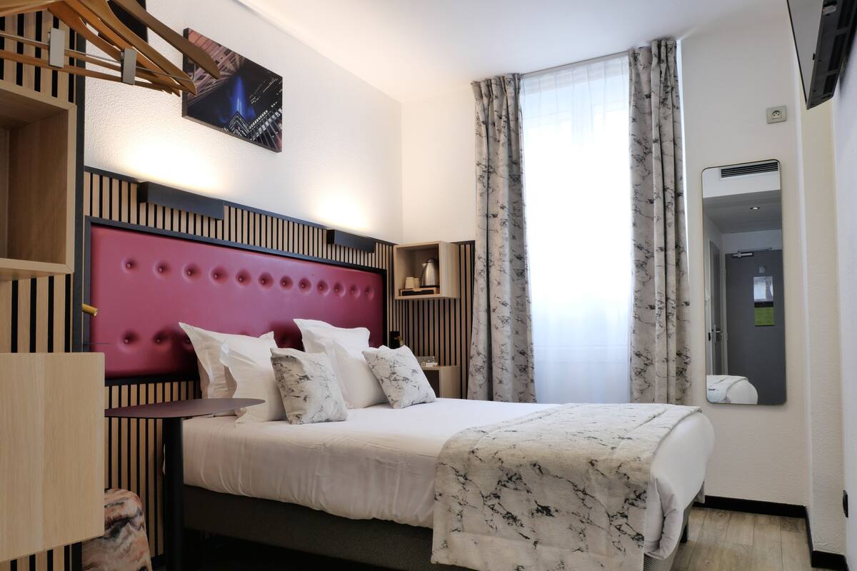 Ein modernes Hotelzimmer mit einem roten, bezogenen Kopfteil, einem Bett mit weißer Bettwäsche, einer Decke mit Marmormuster und einem großen Fenster mit durchsichtigen Vorhängen.