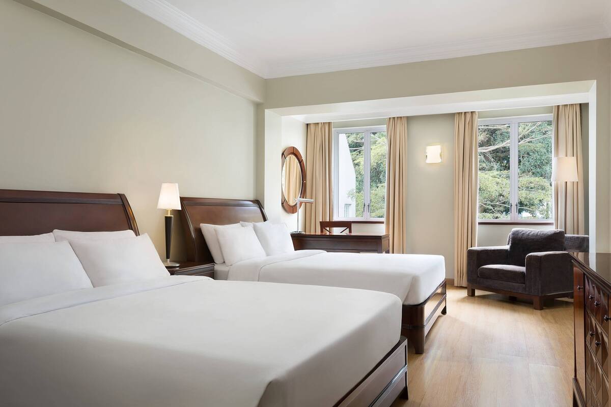 Ein Hotelzimmer mit zwei Betten, einem Sessel, einem Schreibtisch und einem Fenster mit Vorhängen.