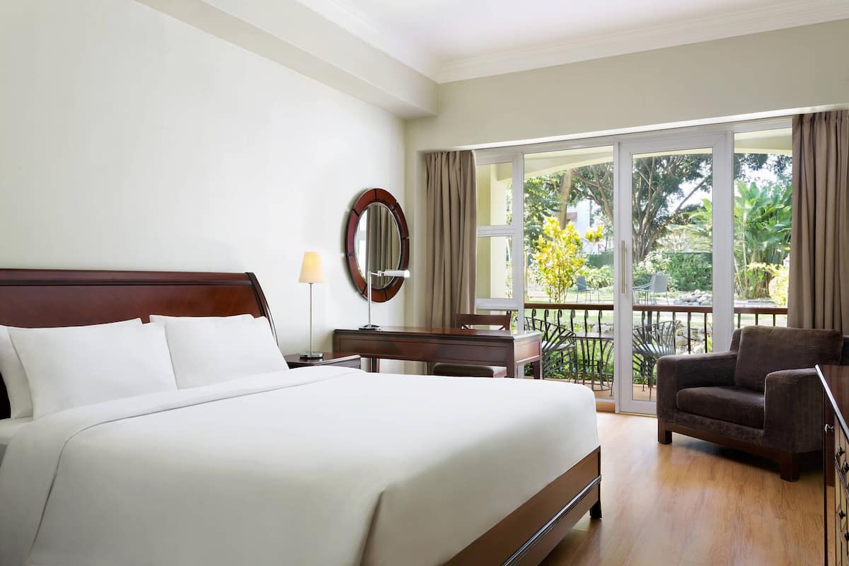 Ein Hotelzimmer mit einem großen Bett, einem Schreibtisch, einem Sessel und einem Balkon mit Blick auf Grünflächen.