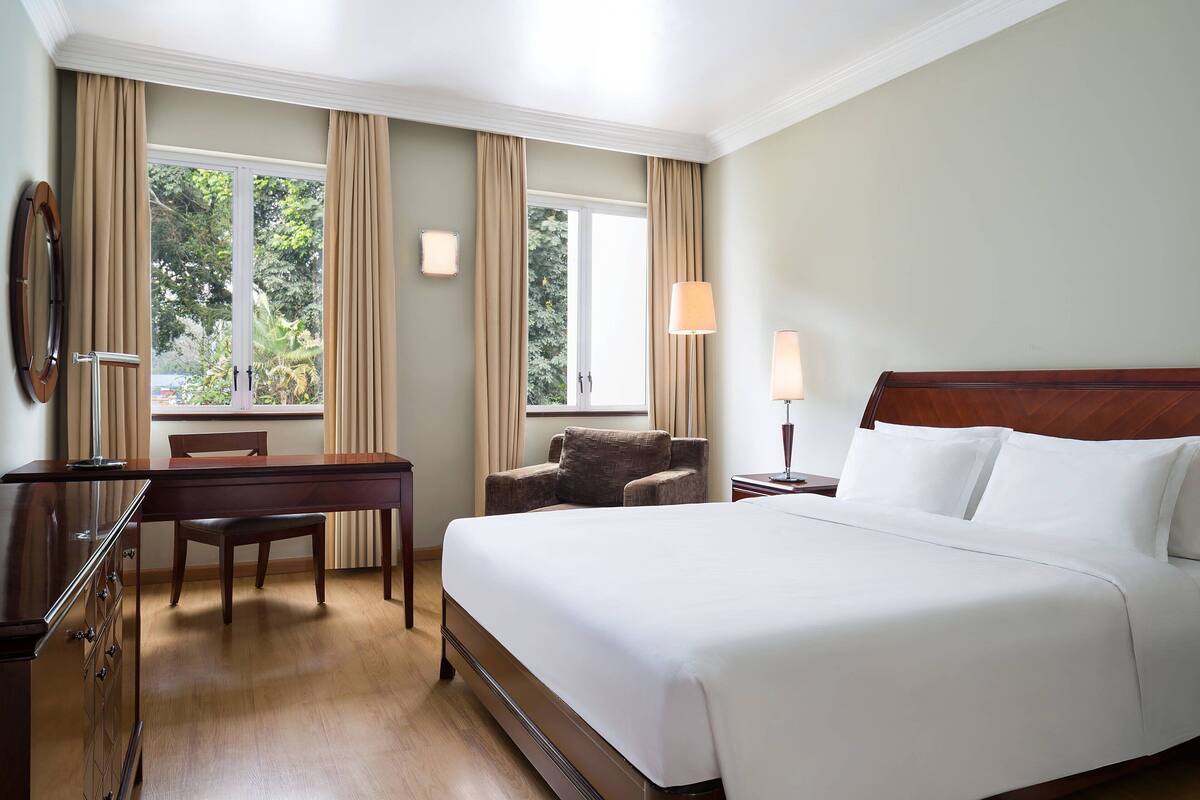 Ein Hotelzimmer mit einem großen Bett, einem Schreibtisch, einem Stuhl und zwei fensterbelaubten Fenstern.