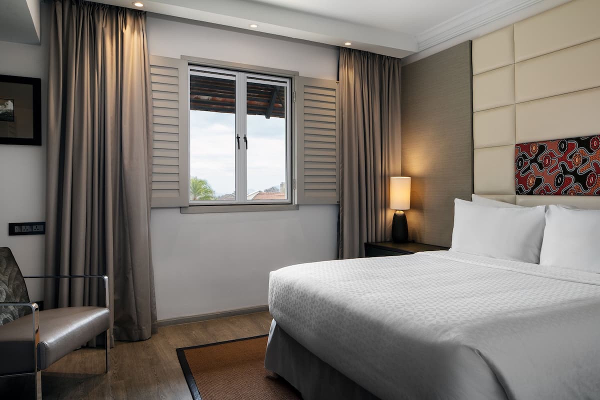 Ein Hotelzimmer mit Bett, Sessel, Fenster mit Rollläden, Lampe und Wandbild mit Muster.