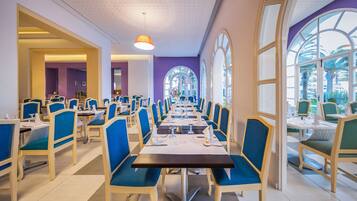Un espace repas avec des tables et des chaises, de grandes fenêtres et un plafond orné d’un motif décoratif.