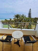 Varanda com mesa e duas cadeiras, vista para uma praia e palmeiras.