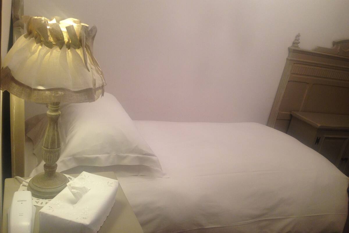 Una cama bien tendida con sábanas blancas, una lámpara de noche y un teléfono sobre la mesita de noche.