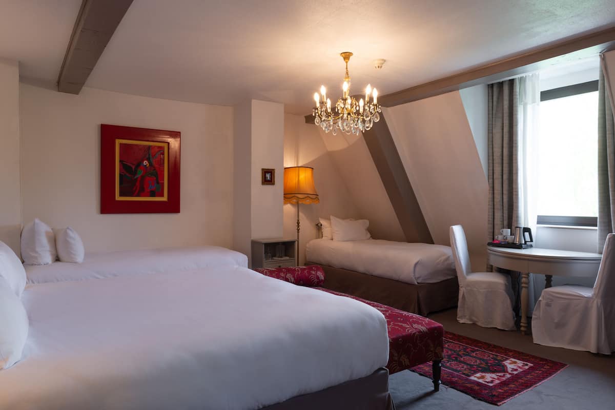 Habitación de hotel con una cama grande, una cama más pequeña, un escritorio con sillas, una araña, un cuadro y una ventana con cortinas.