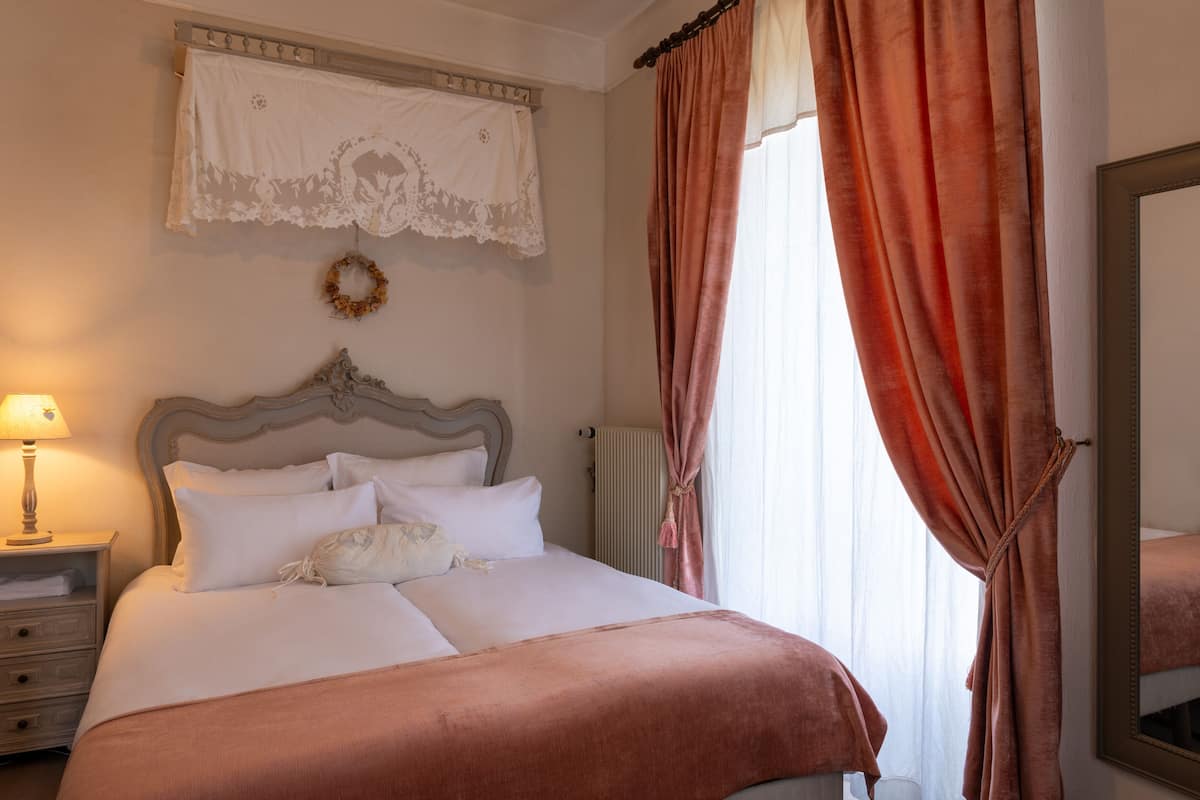 Una cama bien hecha con sábanas blancas y una manta marrón, una mesita de noche de madera con una lámpara, un espejo en la pared y una ventana con cortinas rojas.