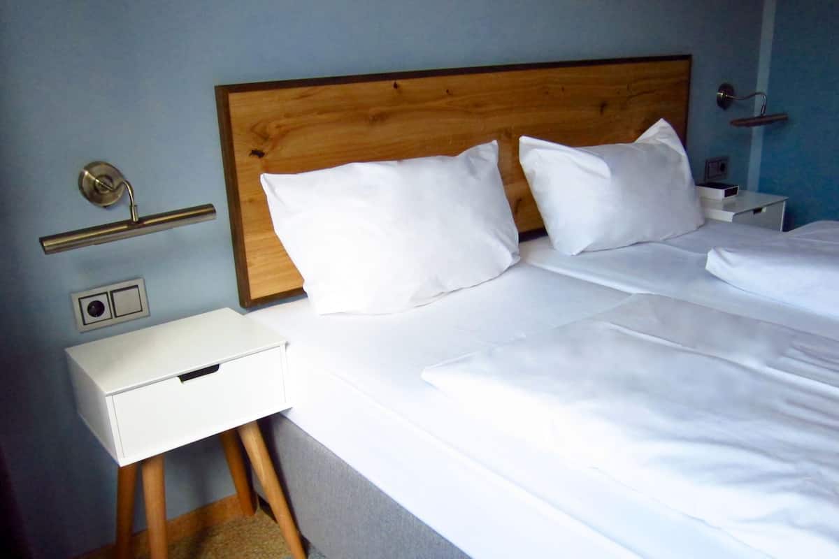 A hotel room with a wooden headboard, white bedding, a bedside table, and a wall-mounted light fixture.