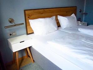 Quarto de hotel com cabeceira de madeira, roupa de cama branca, mesa de cabeceira e luminária na parede.