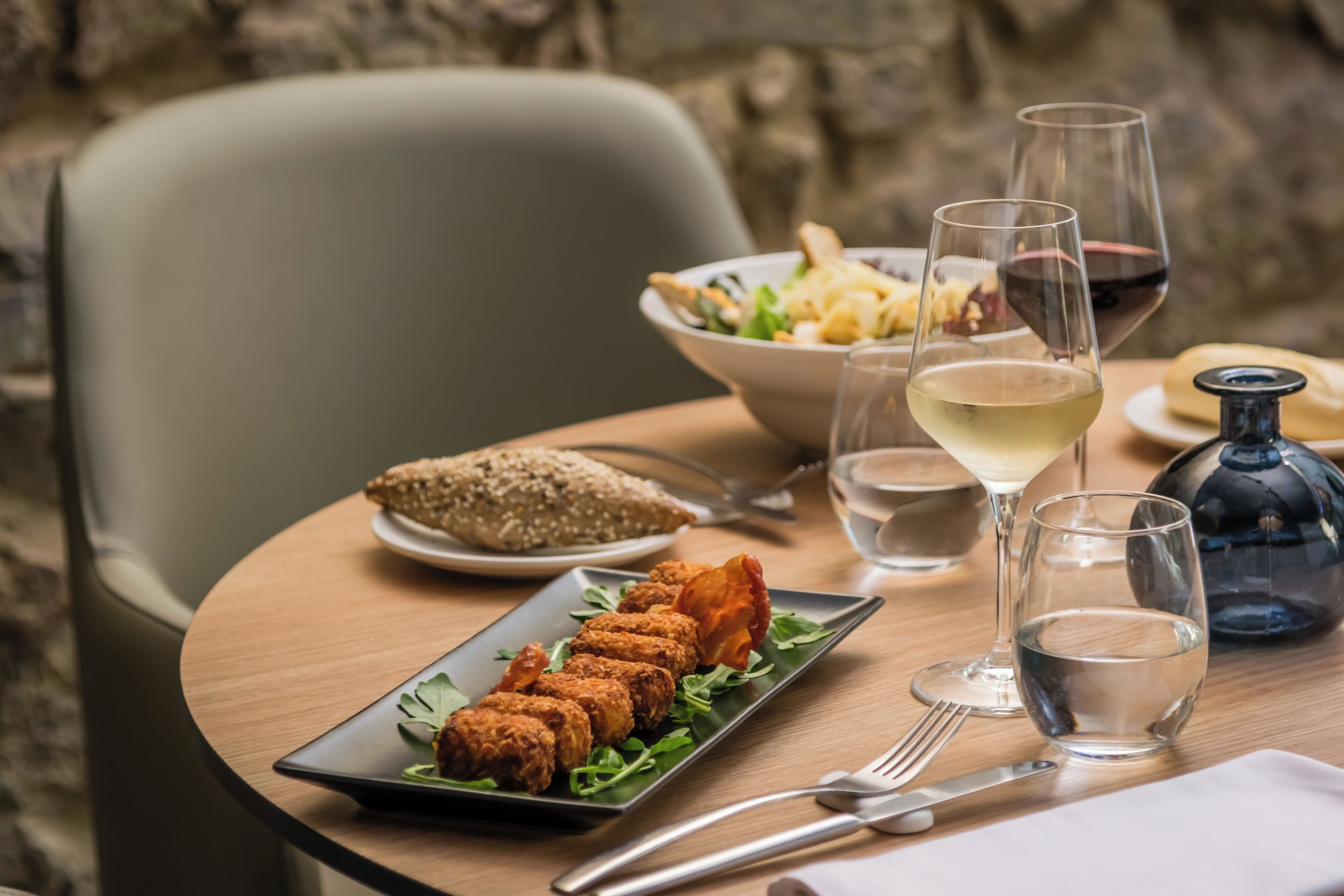 Una mesa de comedor con un plato de comida frita, un bol de ensalada y dos copas de vino.