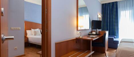 A hotel room with a bed, a desk with a chair, a bench, a mirror, and a TV.