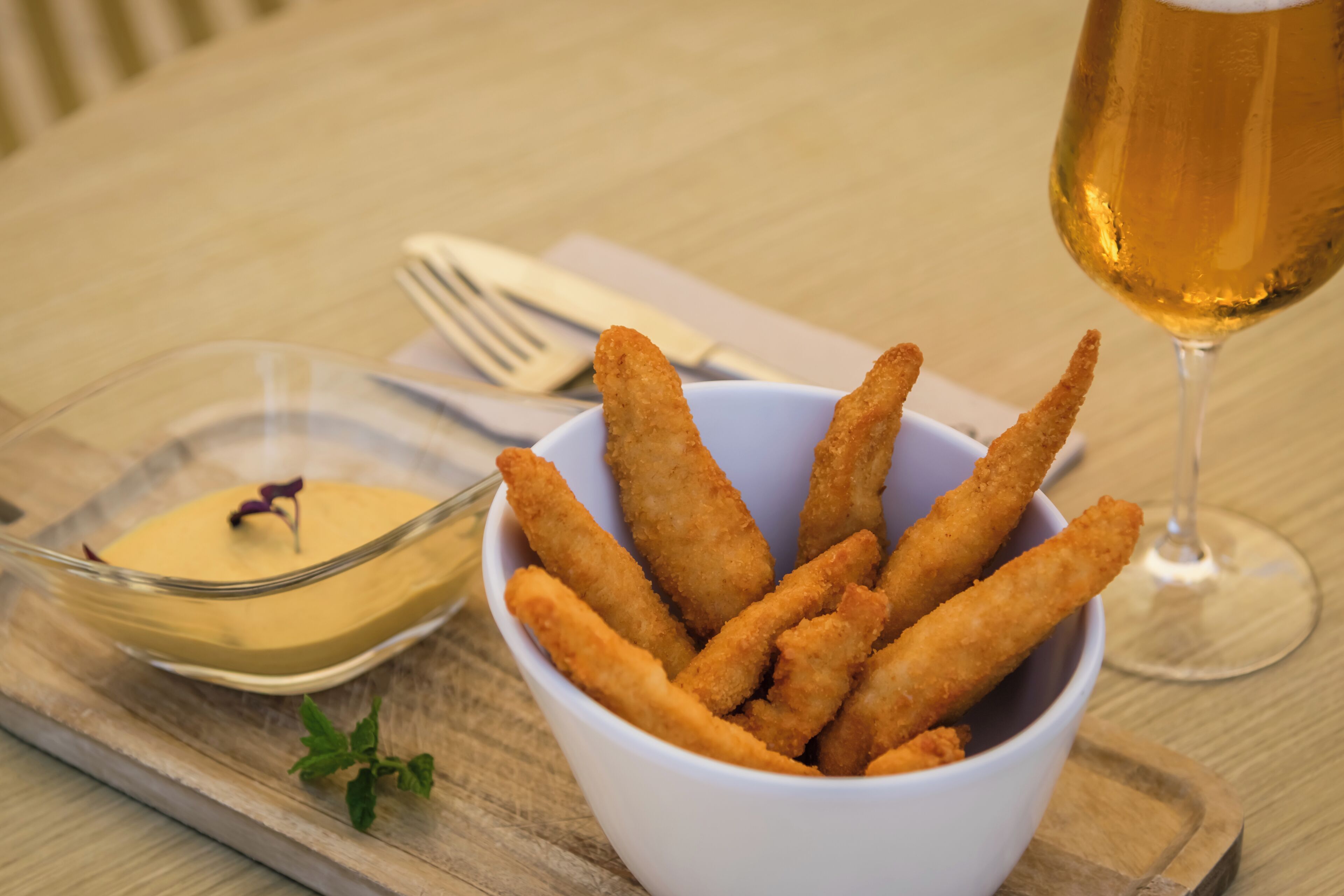 Un bol de alimentos rebozados, una jarra de cerveza y un recipiente pequeño con salsa sobre una bandeja de madera.