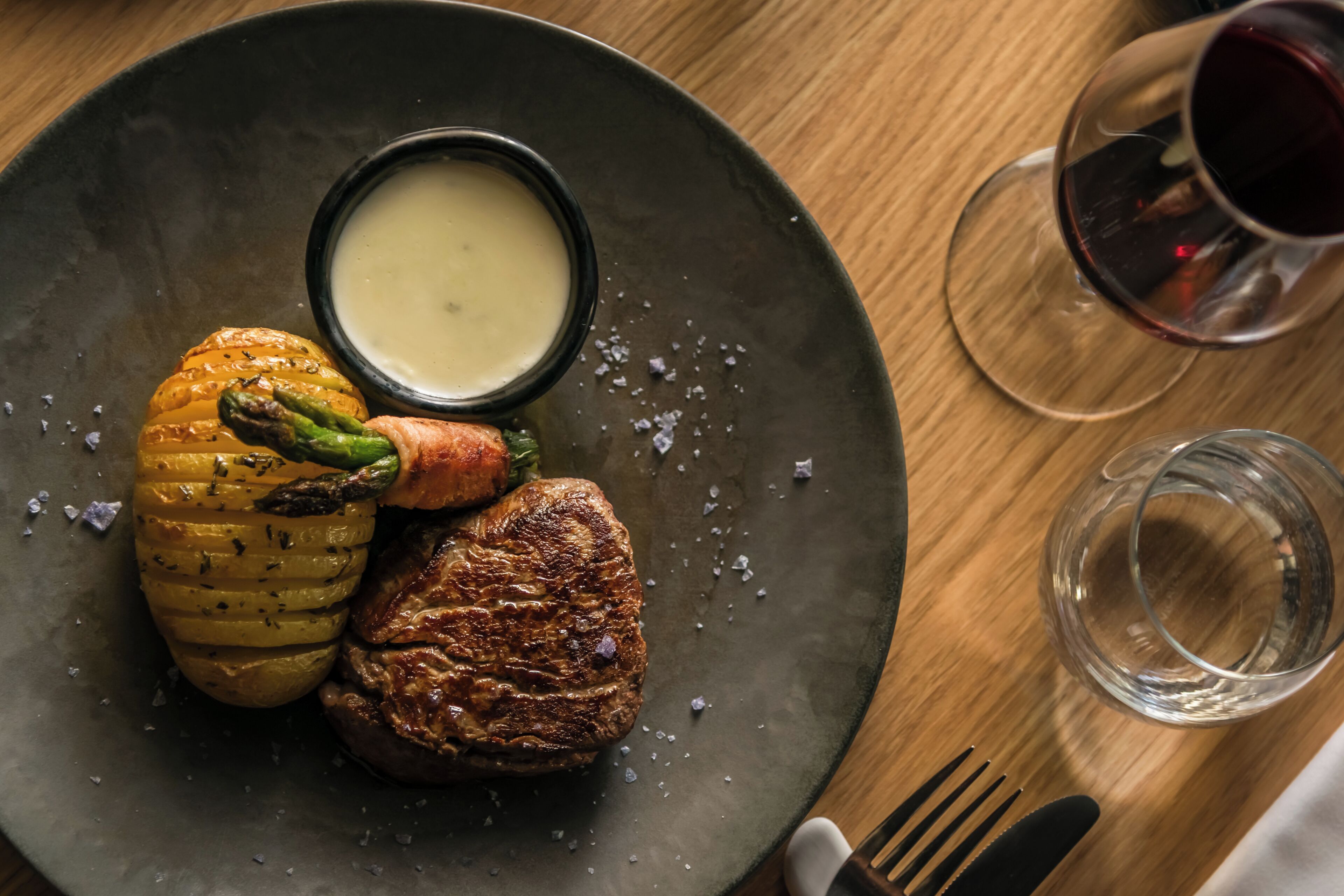 A plate with roasted vegetables and meat, a small bowl of sauce, and a glass of red wine on a wooden table.