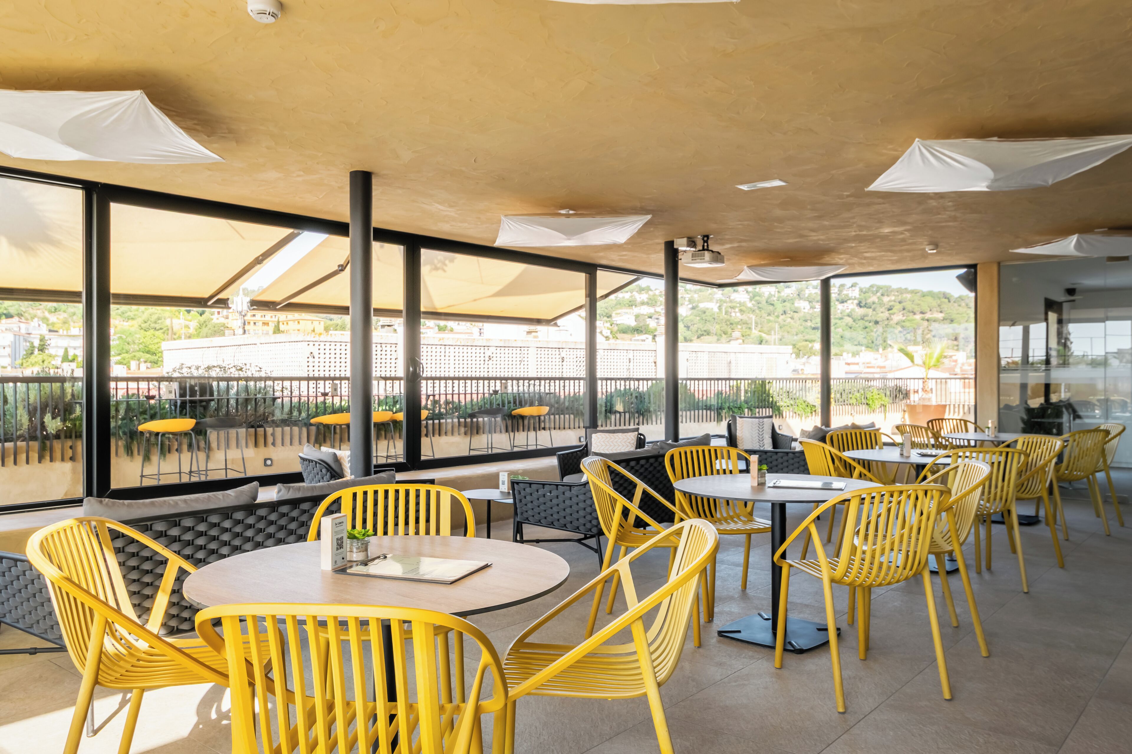 An outdoor dining area with yellow chairs and tables, large windows, and a view of buildings and greenery.