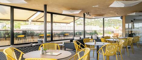 An outdoor dining area with yellow chairs and tables, large windows, and a view of buildings and greenery.