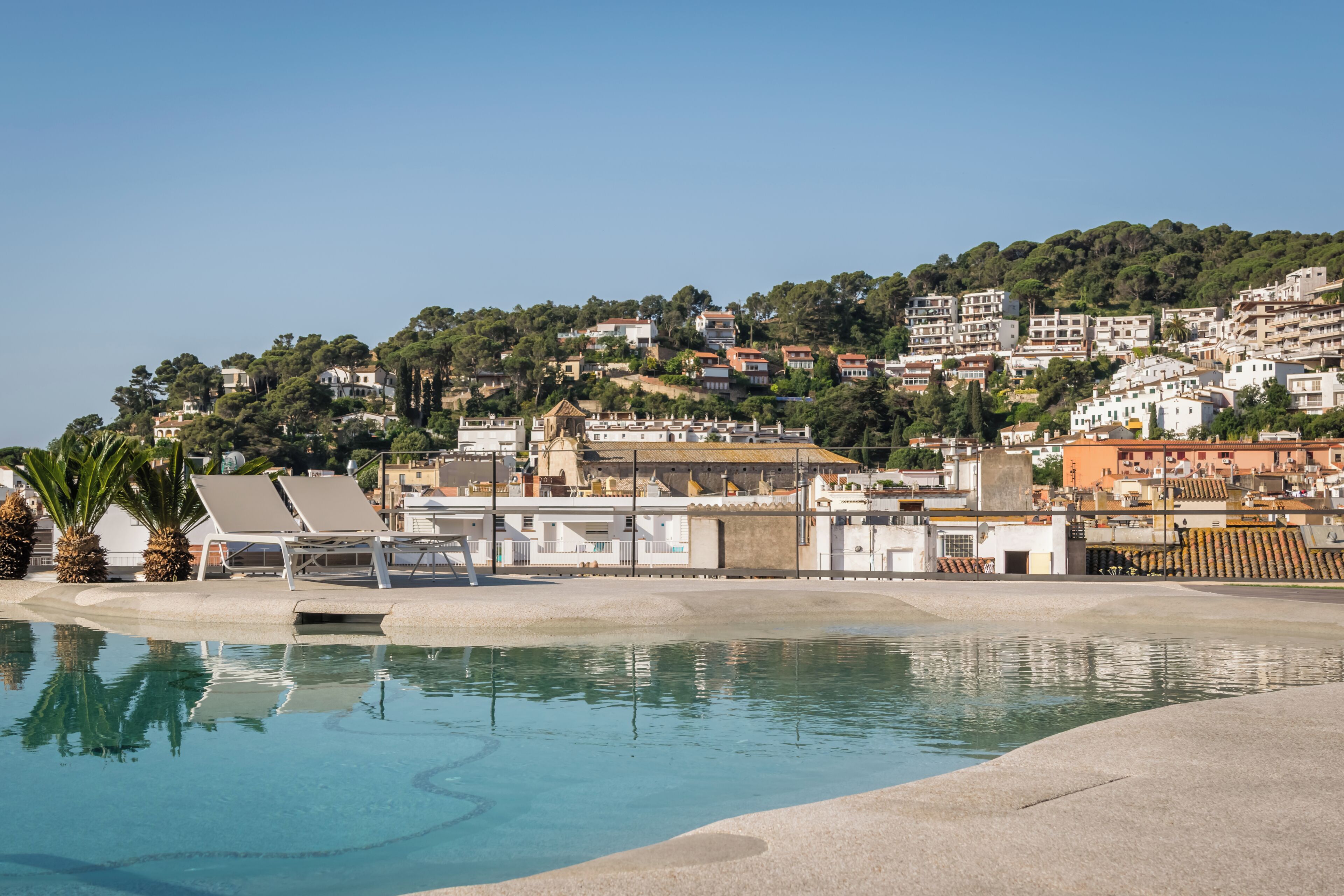 Una piscina con sillas de descanso, un edificio de techo plano y una colina con edificaciones residenciales.
