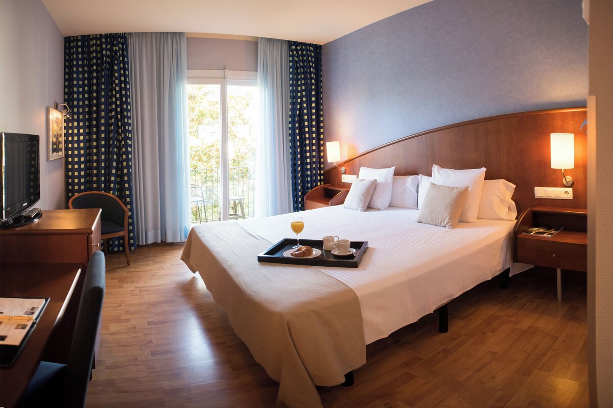 A hotel room with a large bed, a desk with a chair, a TV, and a balcony with curtains.