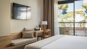 A hotel room with a bed, a desk with a lamp, a TV, a balcony with chairs, and a view of greenery. - Hotel Delfín (Tossa de Mar)