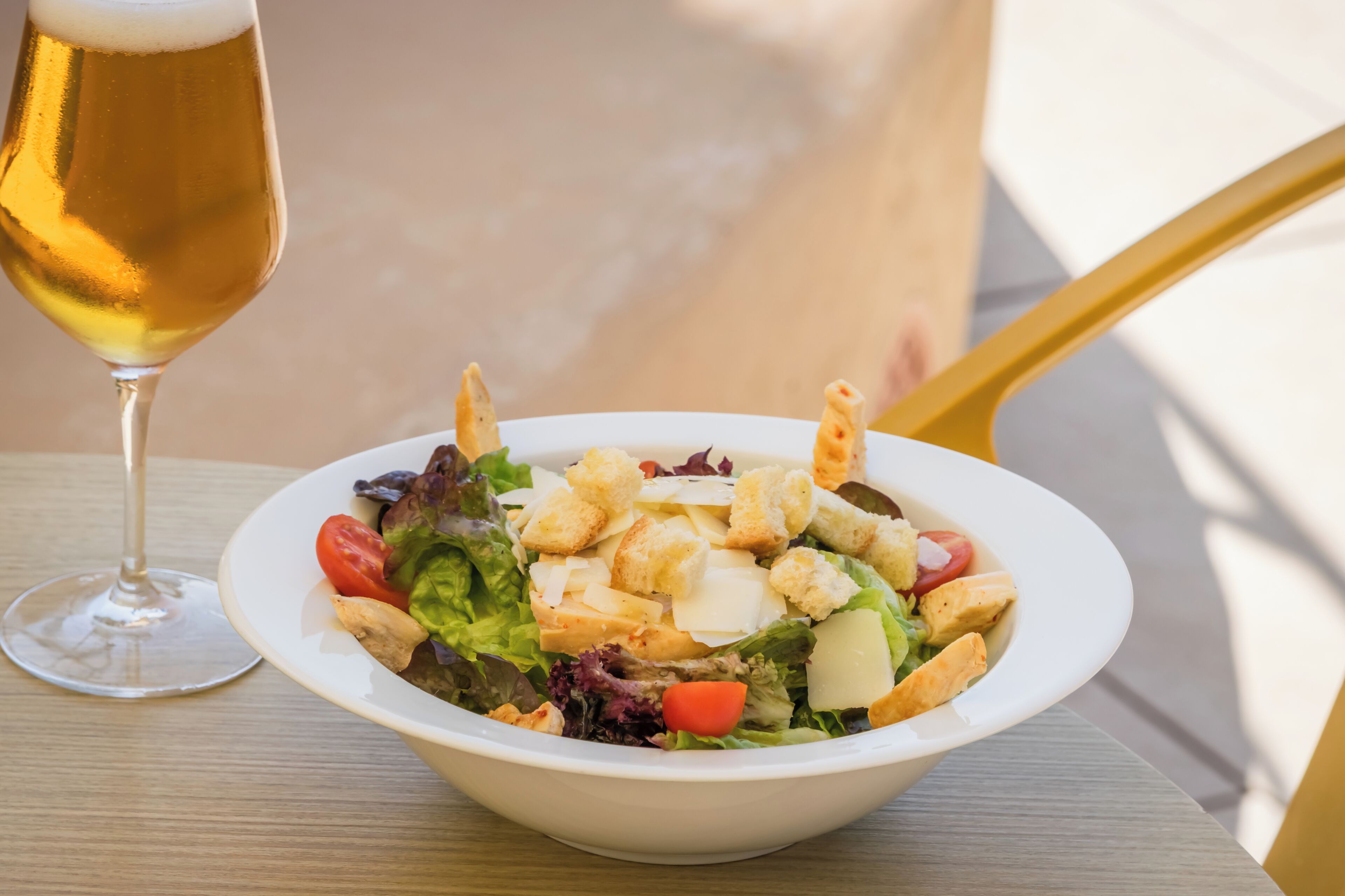 Un tazón blanco con ensalada, crutones y queso, una jarra de cerveza y una silla amarilla.