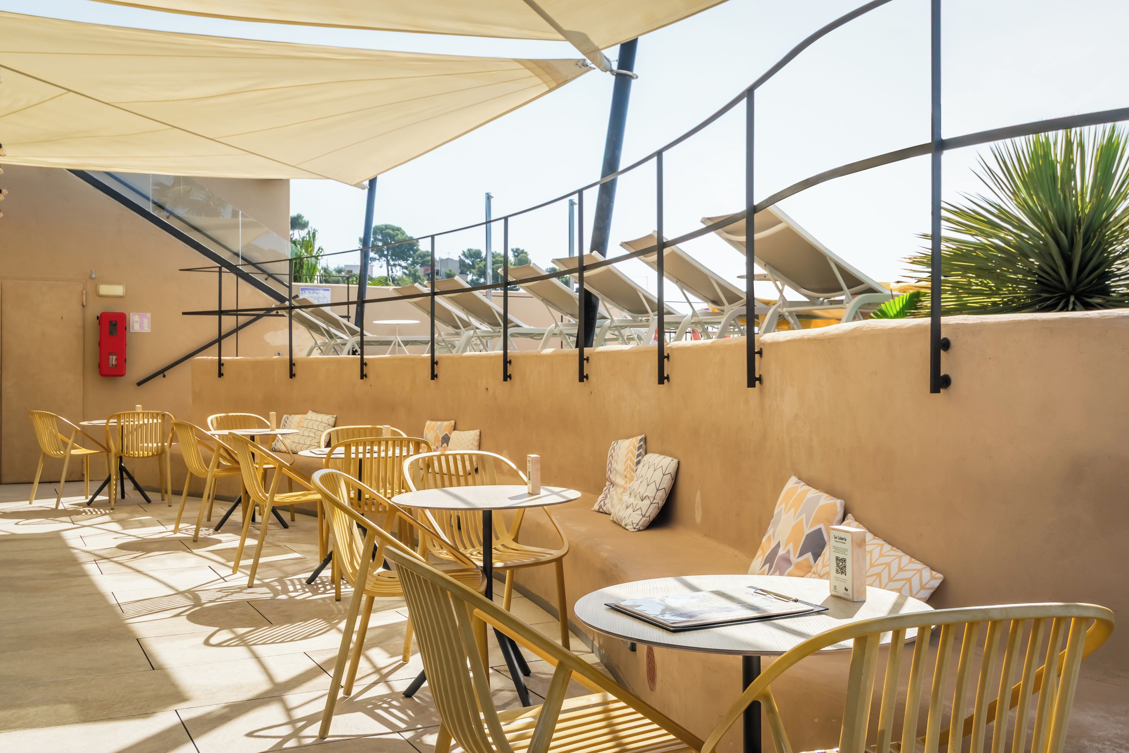An outdoor seating area with yellow chairs and tables, a sofa with patterned cushions, and a large umbrella providing shade.