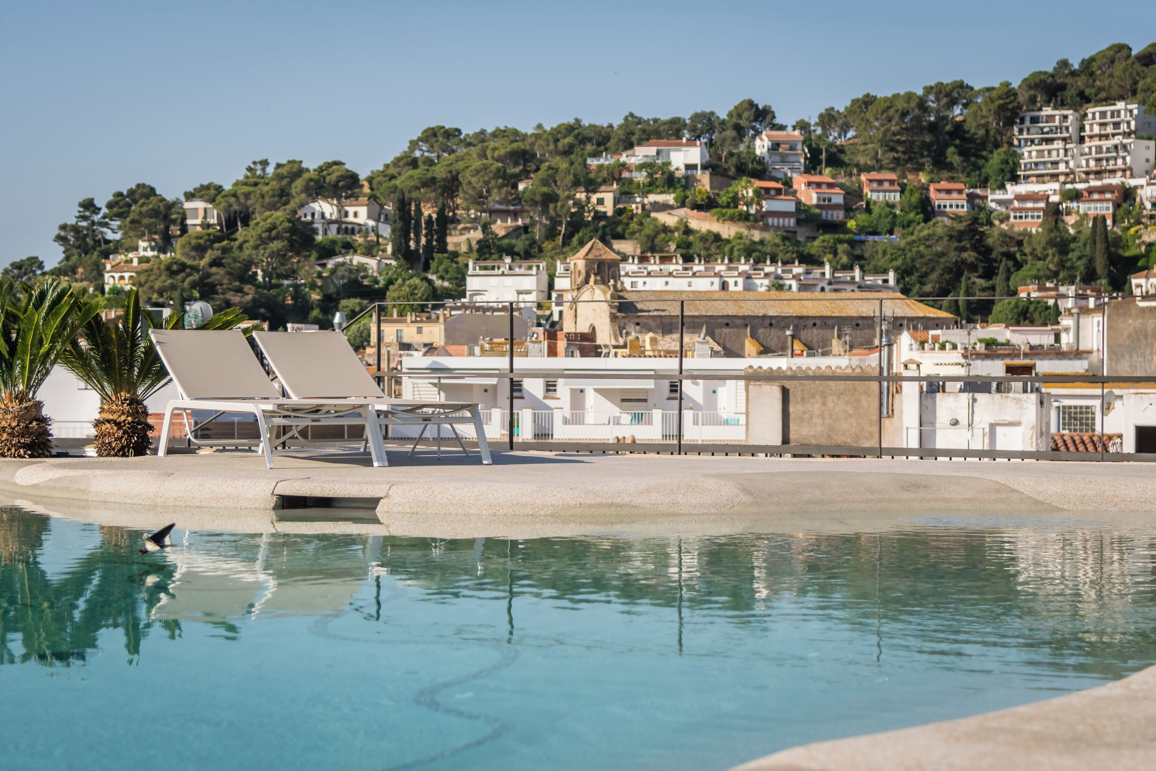 Una piscina con sillones de descanso, una palmera y vistas a una colina con edificios.