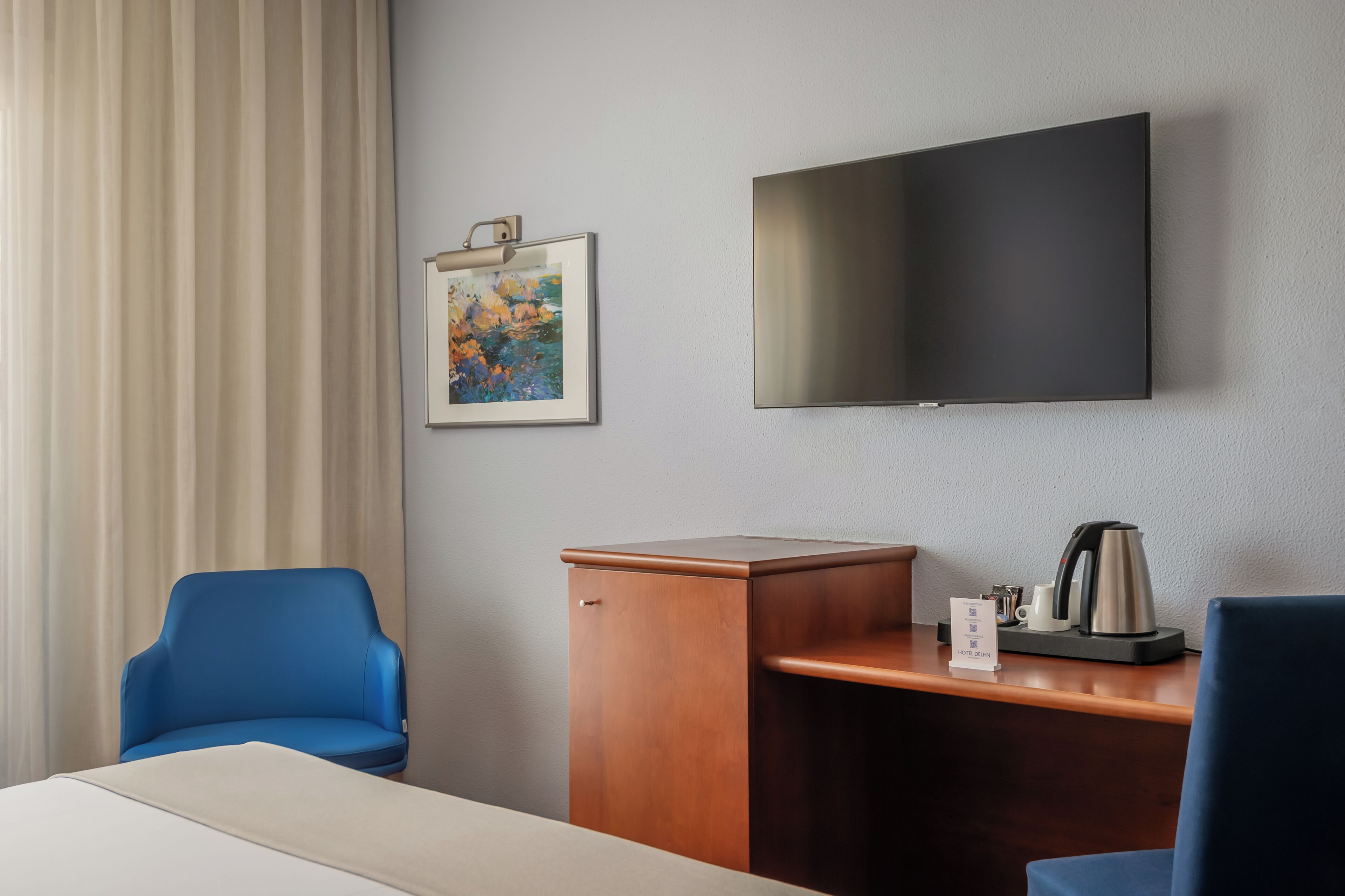 Hotellihuone, jossa on sänky, työpöytä, kahvinkeitin, televisio, sininen tuoli ja taulu seinällä.