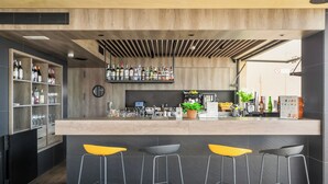A modern bar area with a wooden counter, bar stools, shelves with bottles, and a potted plant. - Hotel Delfín (Tossa de Mar)