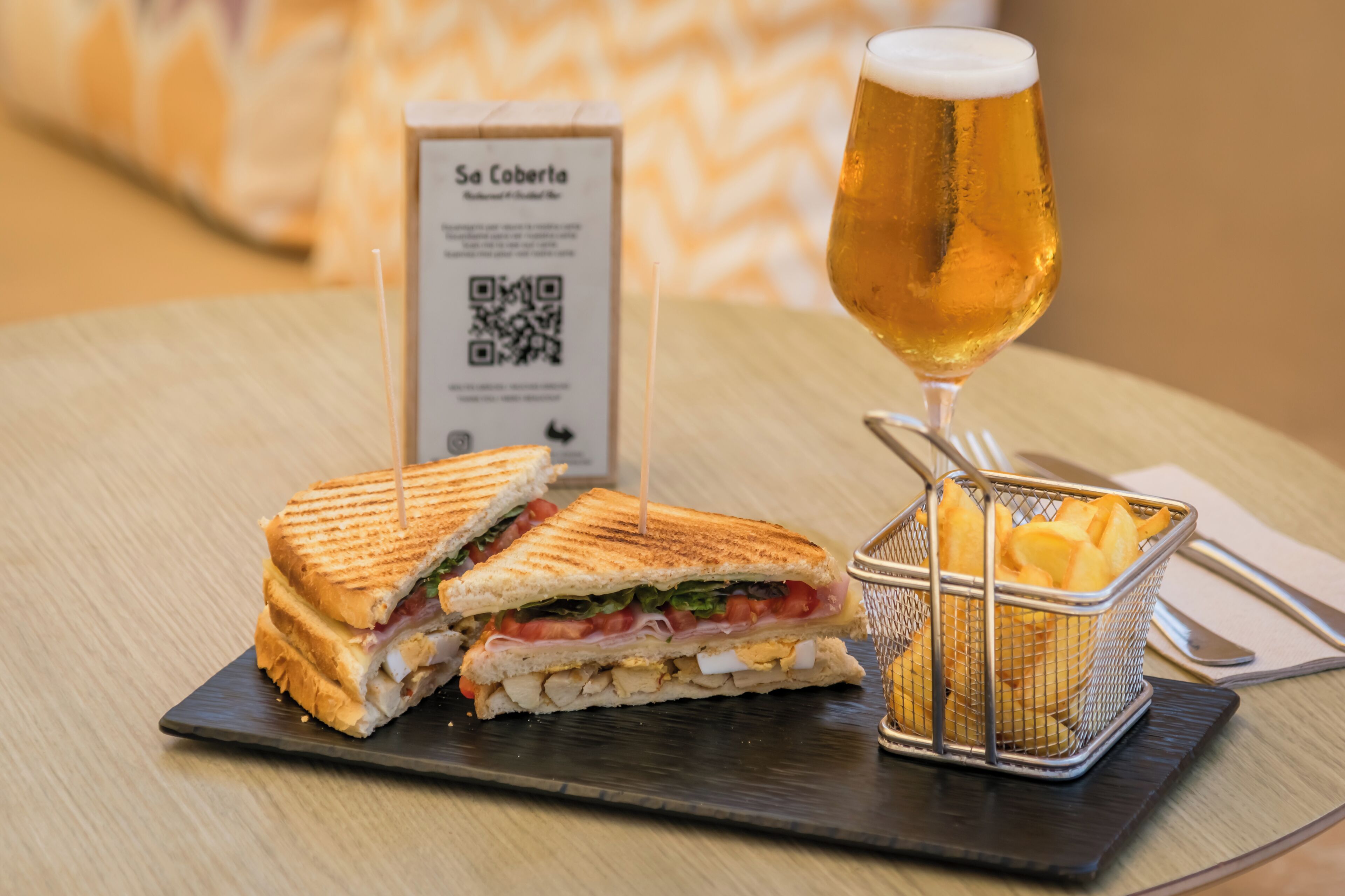 A sandwich cut in half on a dark plate, a glass of beer, and a basket of fries.