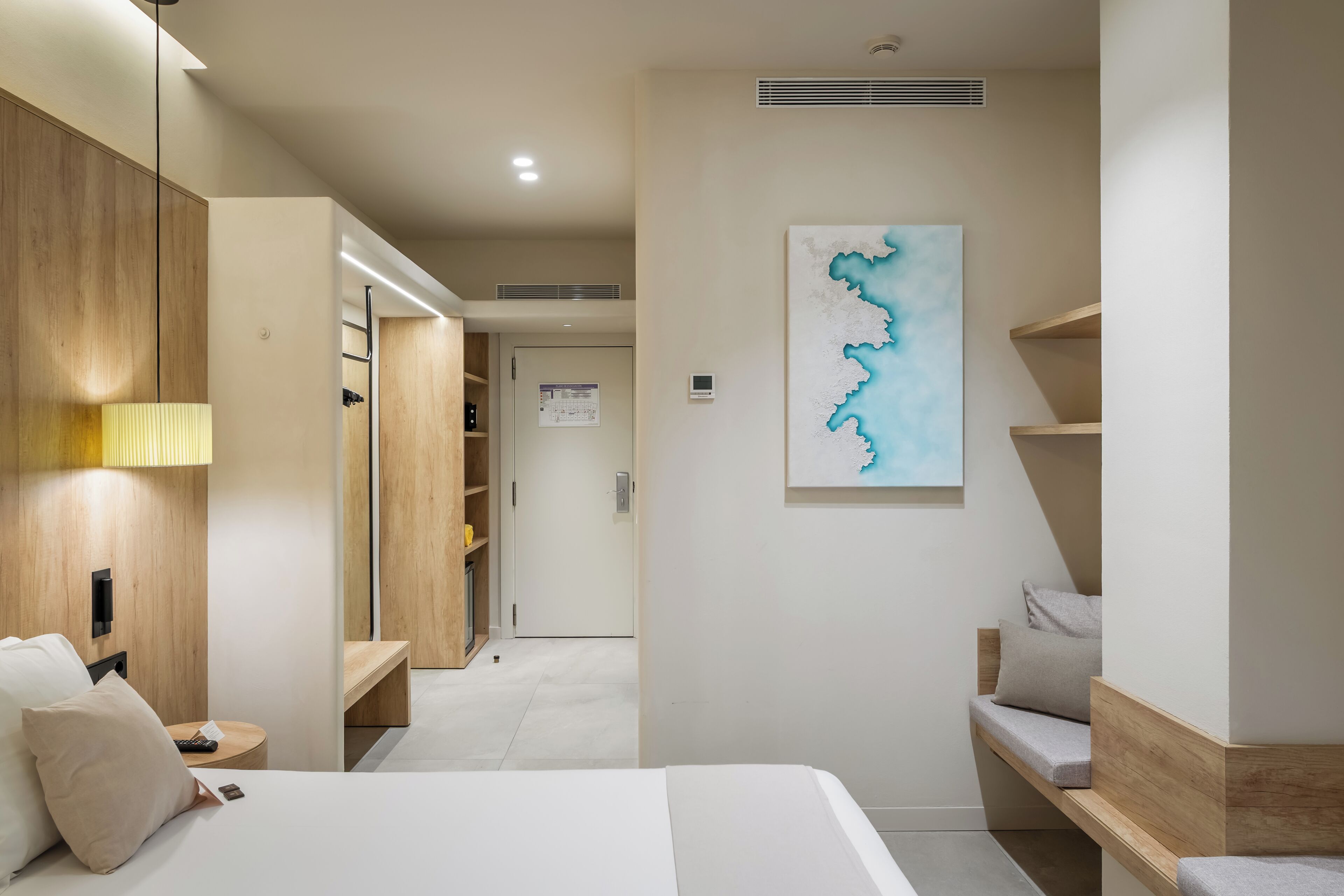A hotel room with a bed, bedside table, lamp, wooden wardrobe, and a wall-mounted artwork.