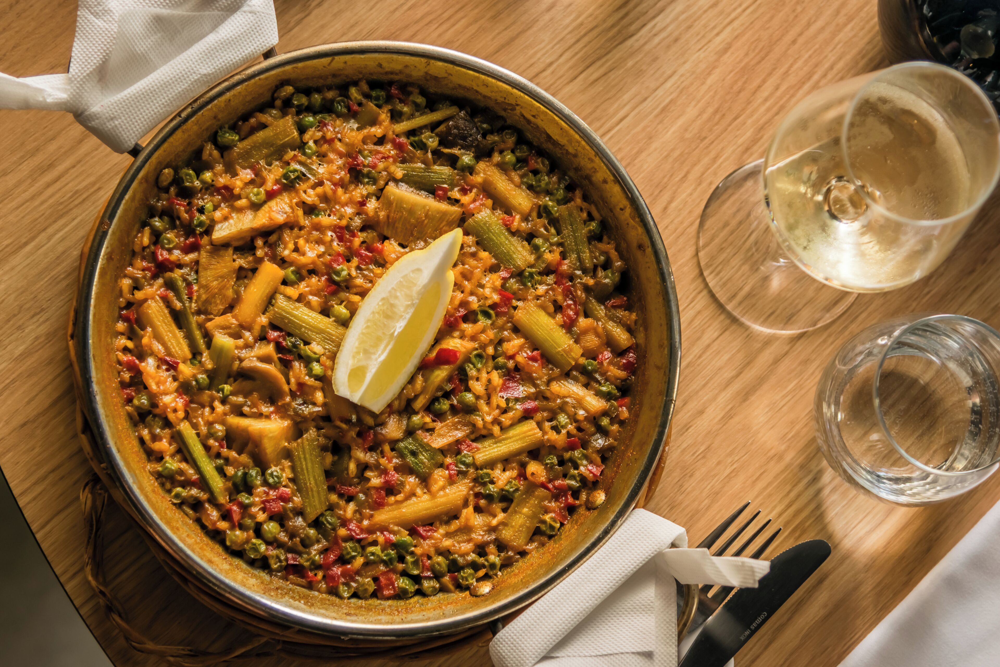 A large pan of vegetable stew with lemon garnish, a glass of white wine, and a fork on a wooden table.