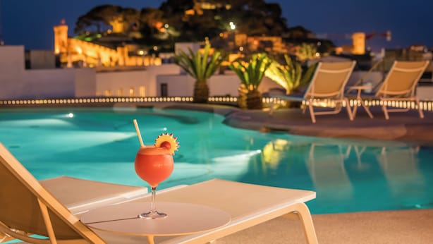 A poolside lounge with a drink on a chair, clear water, and a distant illuminated building. - Hotel Delfín (Tossa de Mar)