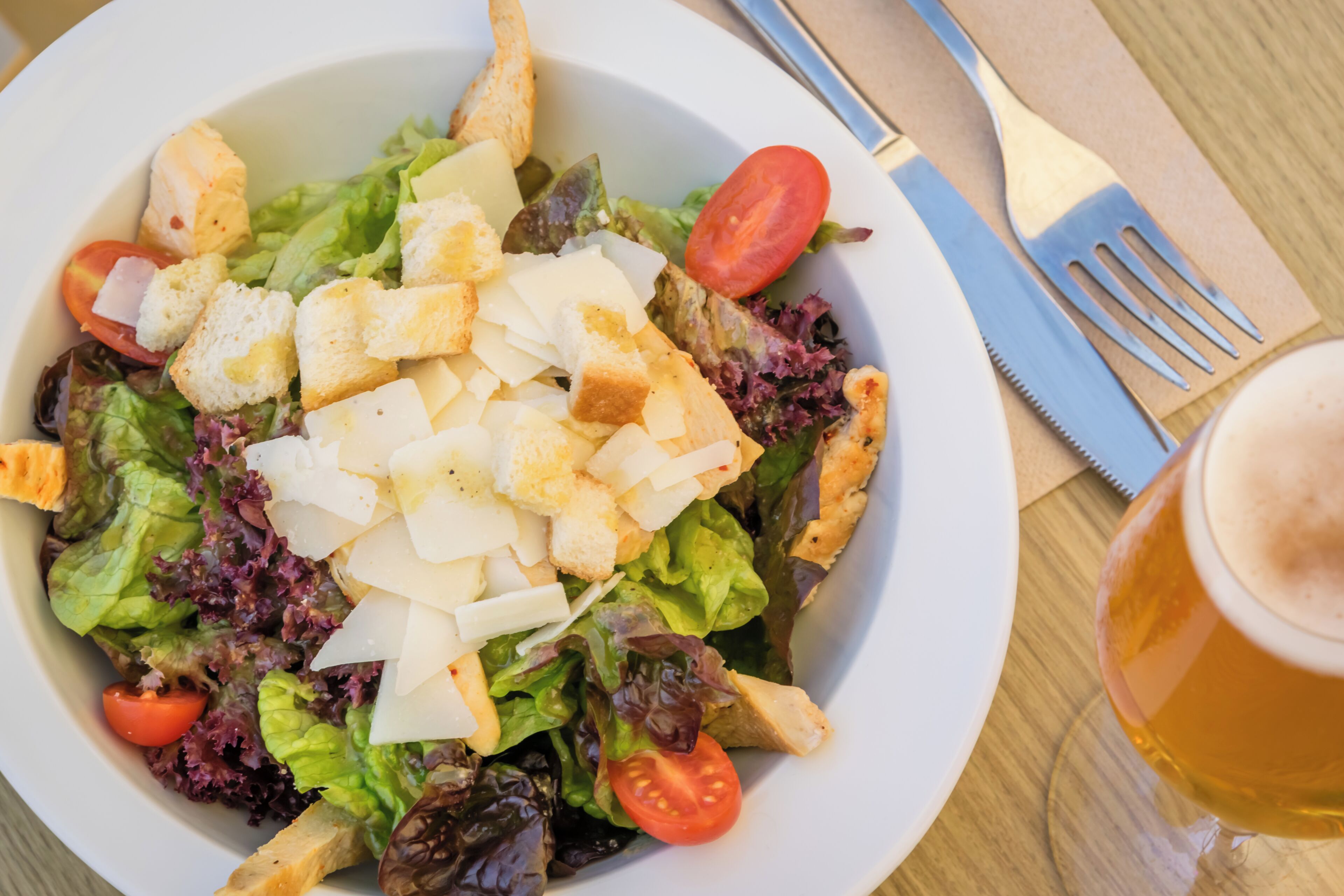 A bowl of salad with croutons and cheese, a glass of beer, and cutlery on a table.