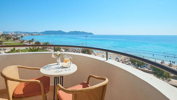 Un balcon avec des fauteuils en osier et une table, donnant sur une plage animée et un ciel bleu dégagé.