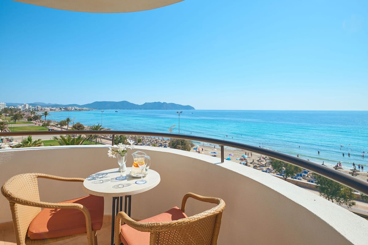 Ein Balkon mit Korbstühlen und einem Tisch, der Blick auf einen Strand mit Menschen und einem klaren, blauen Himmel bietet.