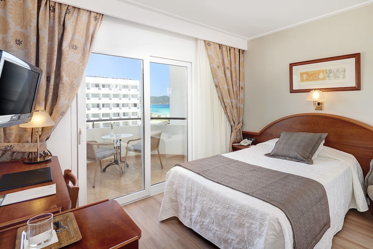 Ein Hotelzimmer mit Bett, Fernseher, Schreibtisch mit Stuhl, einem Glastisch mit Glas und Buch sowie Meerblick durch eine Schiebetür.
