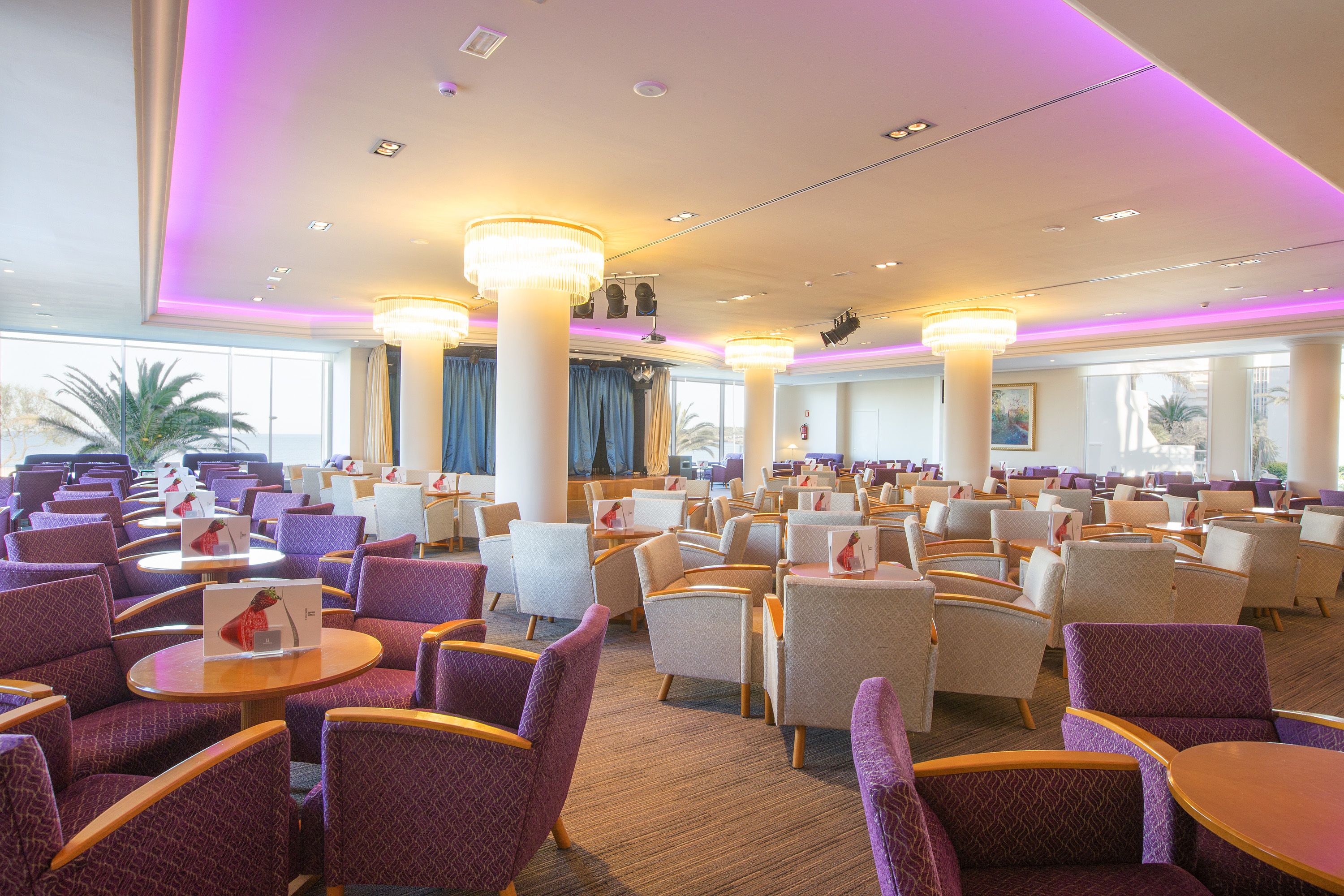 A spacious hotel lobby with purple upholstered chairs and round tables, large pillars, and a view of palm trees through the windows.