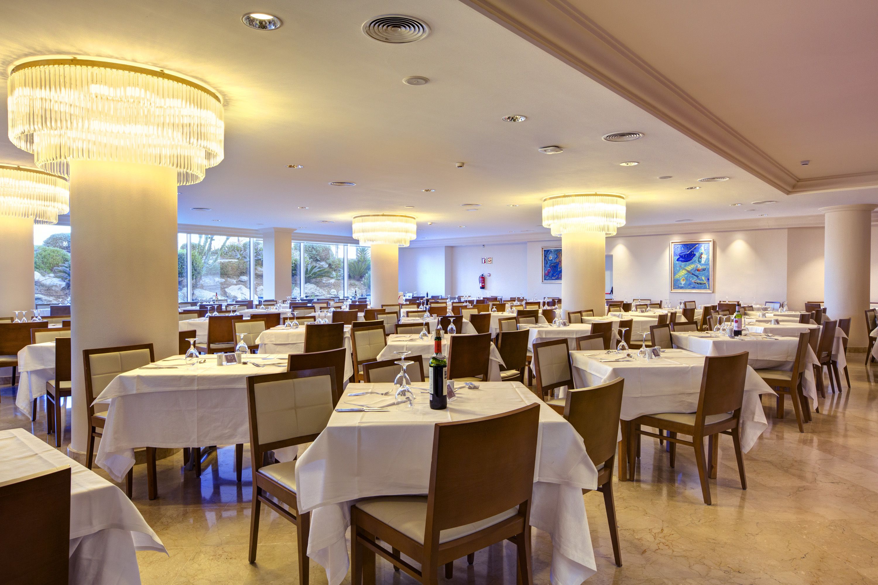 A large dining area with multiple tables and chairs, large chandeliers, and a view of the outdoors.