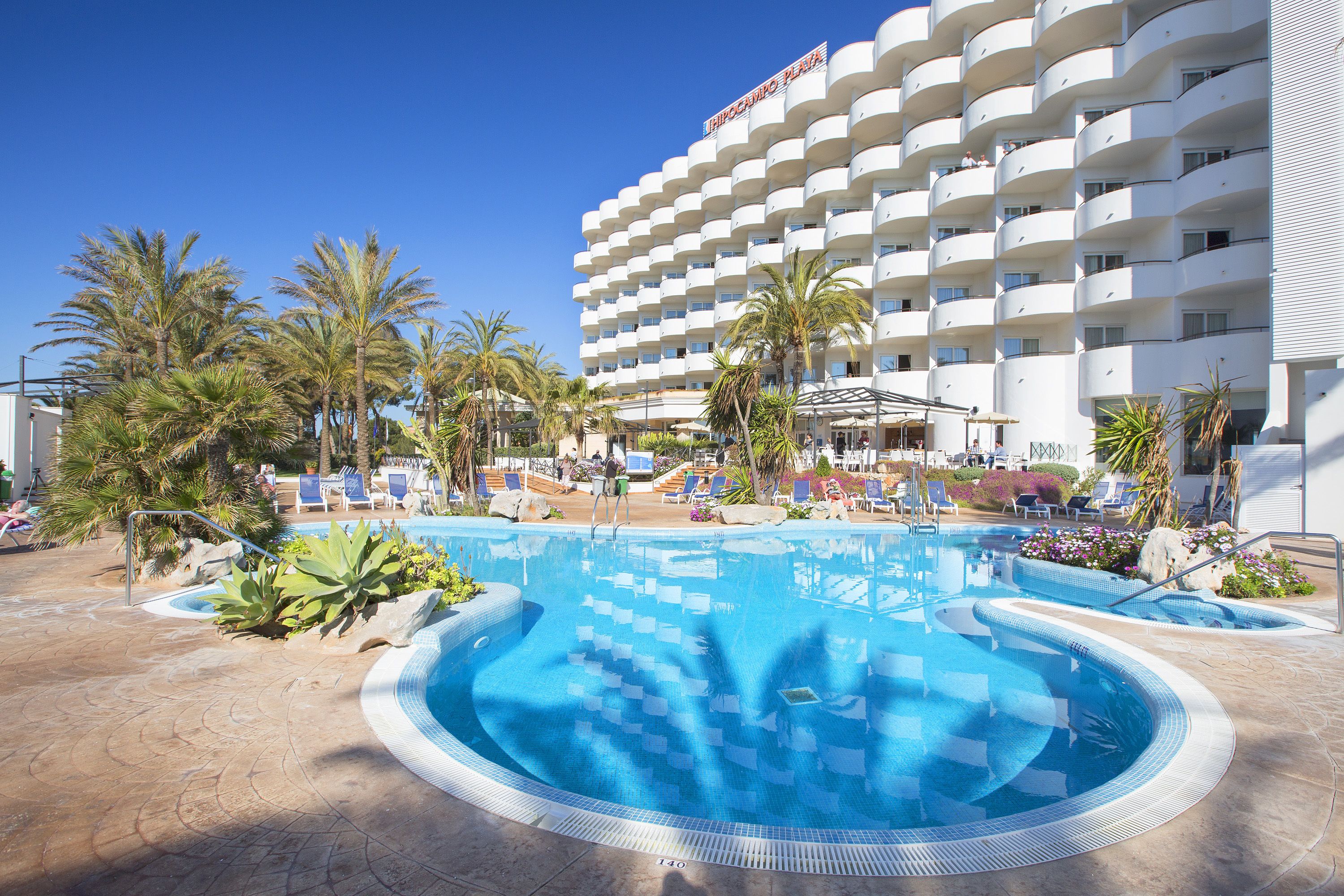 A hotel with a large pool area, palm trees, and a clear blue sky.