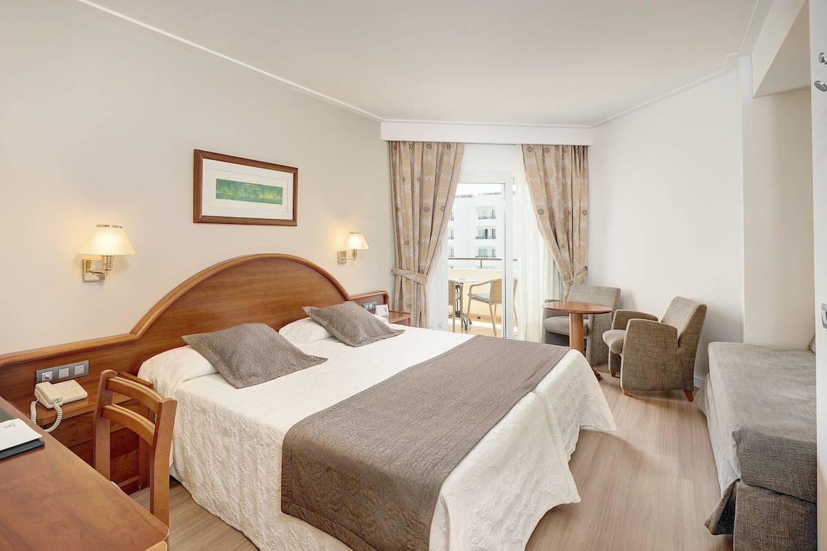 Ein Hotelzimmer mit Bett, Nachttischlampen, Schreibtisch mit Telefon, Stuhl, kleinem Tisch und Fernseher.