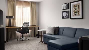 A modern hotel room with a blue sofa, a desk with a chair, a TV, and three framed artworks on the wall.