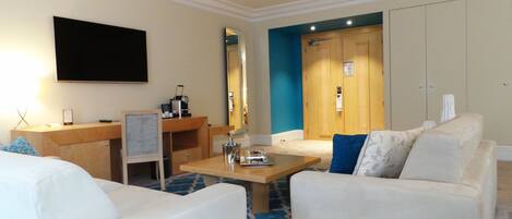 A hotel room with a flat-screen TV, a desk with a coffee maker, a seating area with a sofa and armchairs, and a large wardrobe.