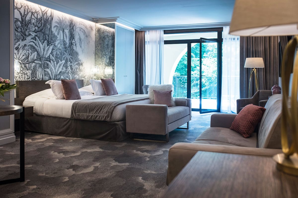 A hotel room with a bed, a sofa, a desk, and a large window with a view of greenery.