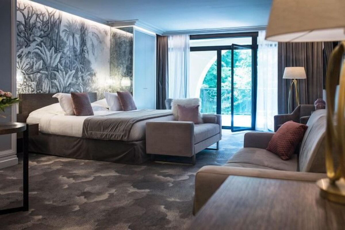 A modern hotel room with a large bed, a sofa, a desk, and a view of greenery through the window.