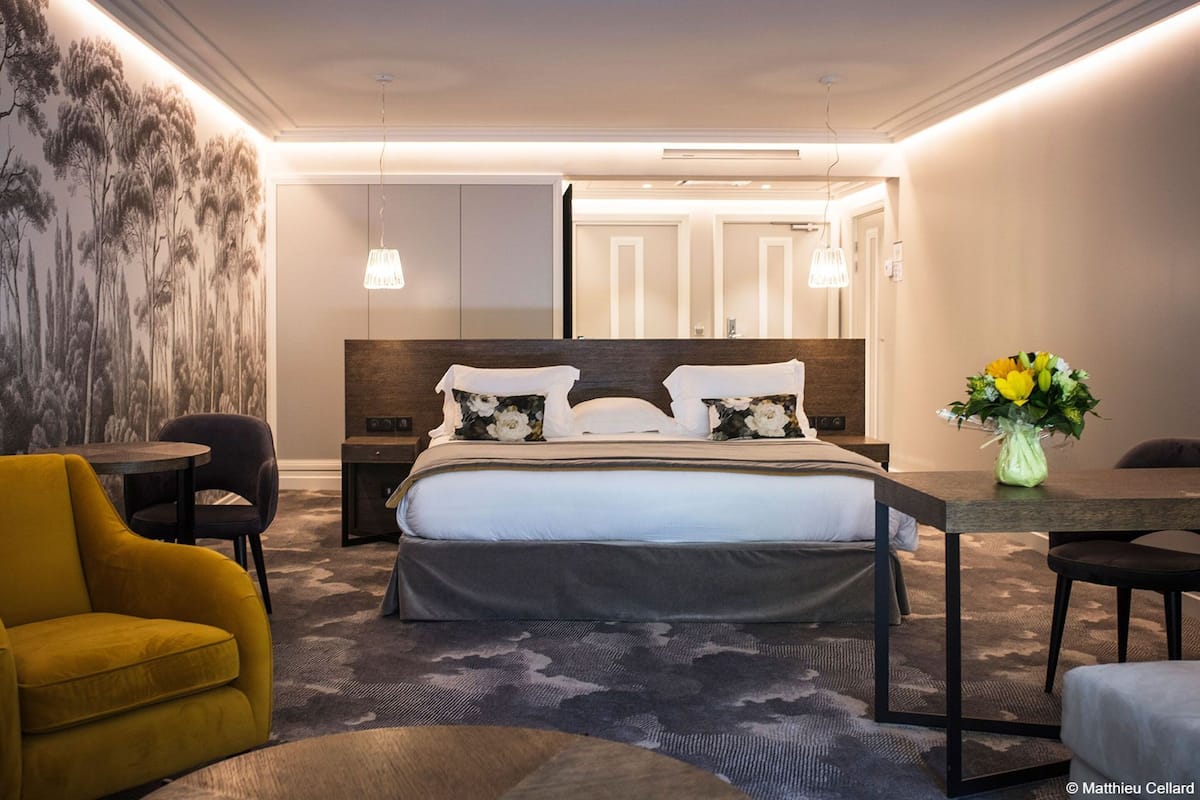 A modern hotel room with a large bed, a desk, a yellow armchair, and a vase of flowers.